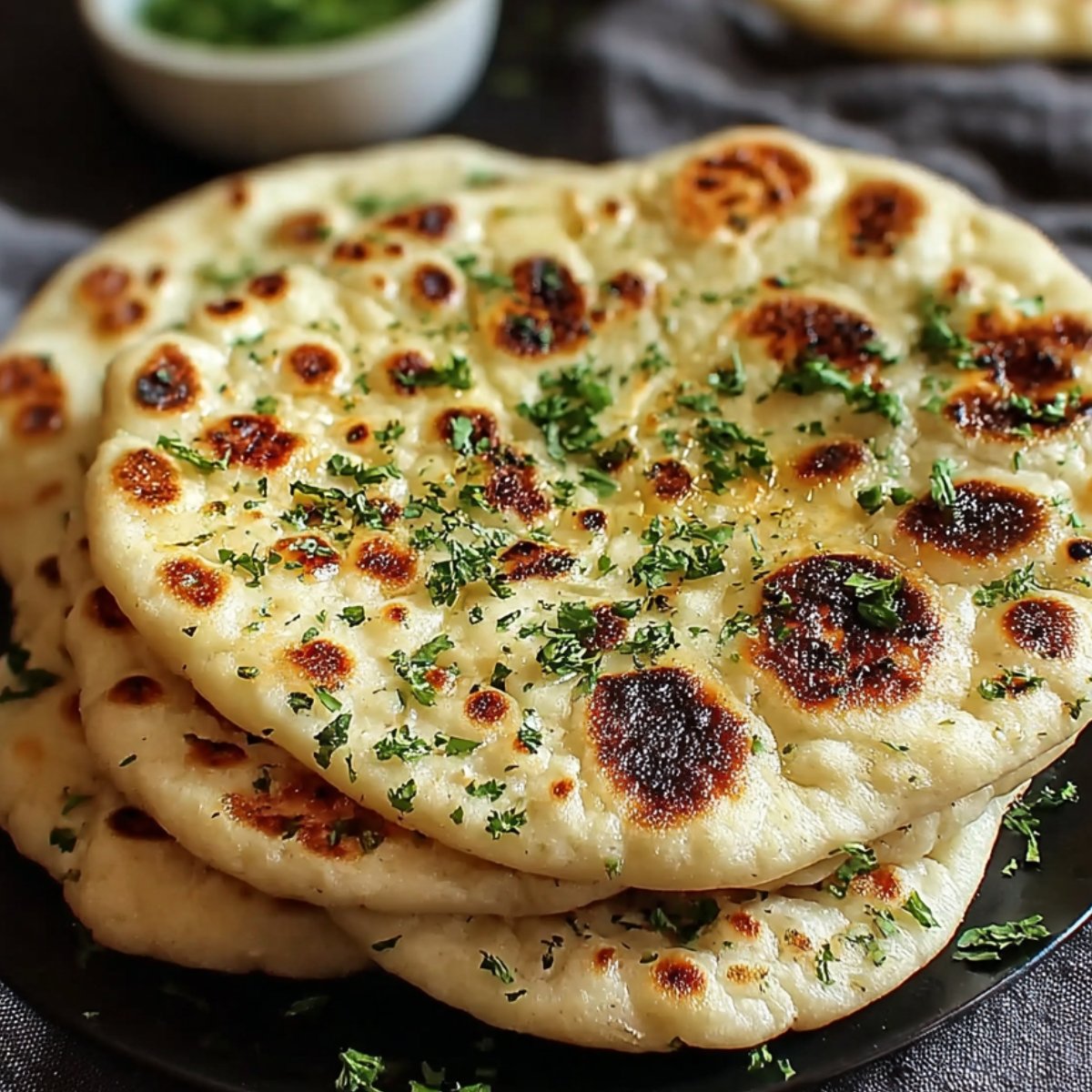 Close‑up of three golden‑brown skillet‑cooked naan bread recipe stacked on a black plate, each brushed with butter, sprinkled with chopped cilantro and nigella seeds, against a dark cloth background.