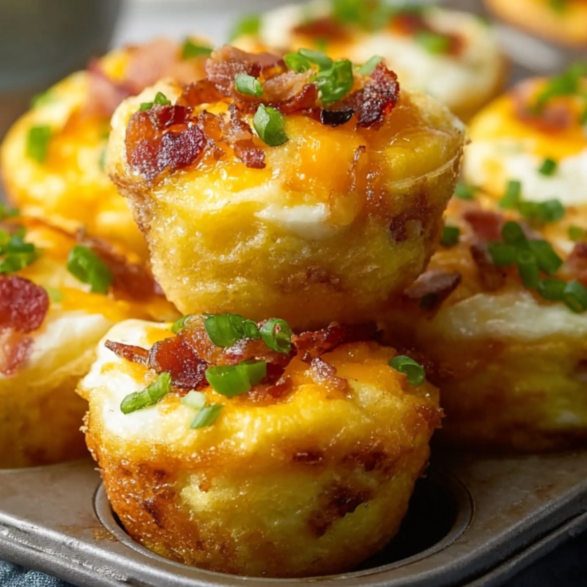 A close-up of three golden Egg Bites Muffin Tins in a dark muffin tin, each topped with melted cheddar, crispy bacon bits, and chopped green onions, with more egg muffins blurred in the background