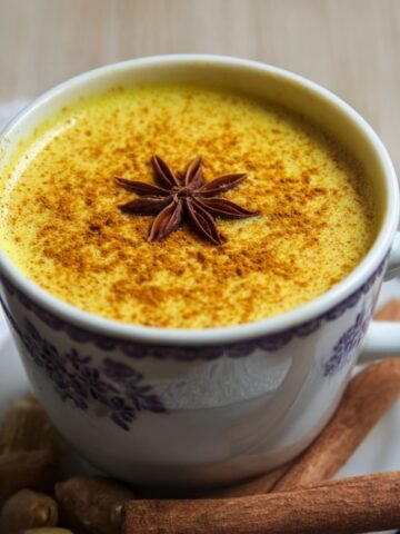 Close‑up three‑quarter view of a white porcelain cup filled with golden milk recipe , sprinkled with turmeric and topped with a star anise. The cup sits on a matching saucer alongside cinnamon sticks and whole nutmeg on a light wooden surface.