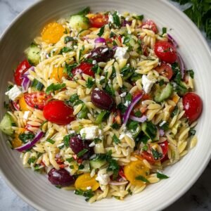 Close‑up overhead shot of finished Greek orzo salad in a white bowl, showing tender orzo mixed with cherry tomatoes, cucumber, red onion, parsley, Kalamata olives, and crumbled feta, all lightly coated in lemon‑olive oil dressing.”