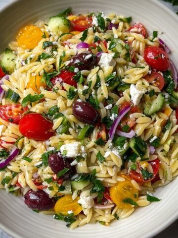 Close‑up overhead shot of finished Greek orzo salad in a white bowl, showing tender orzo mixed with cherry tomatoes, cucumber, red onion, parsley, Kalamata olives, and crumbled feta, all lightly coated in lemon‑olive oil dressing.”