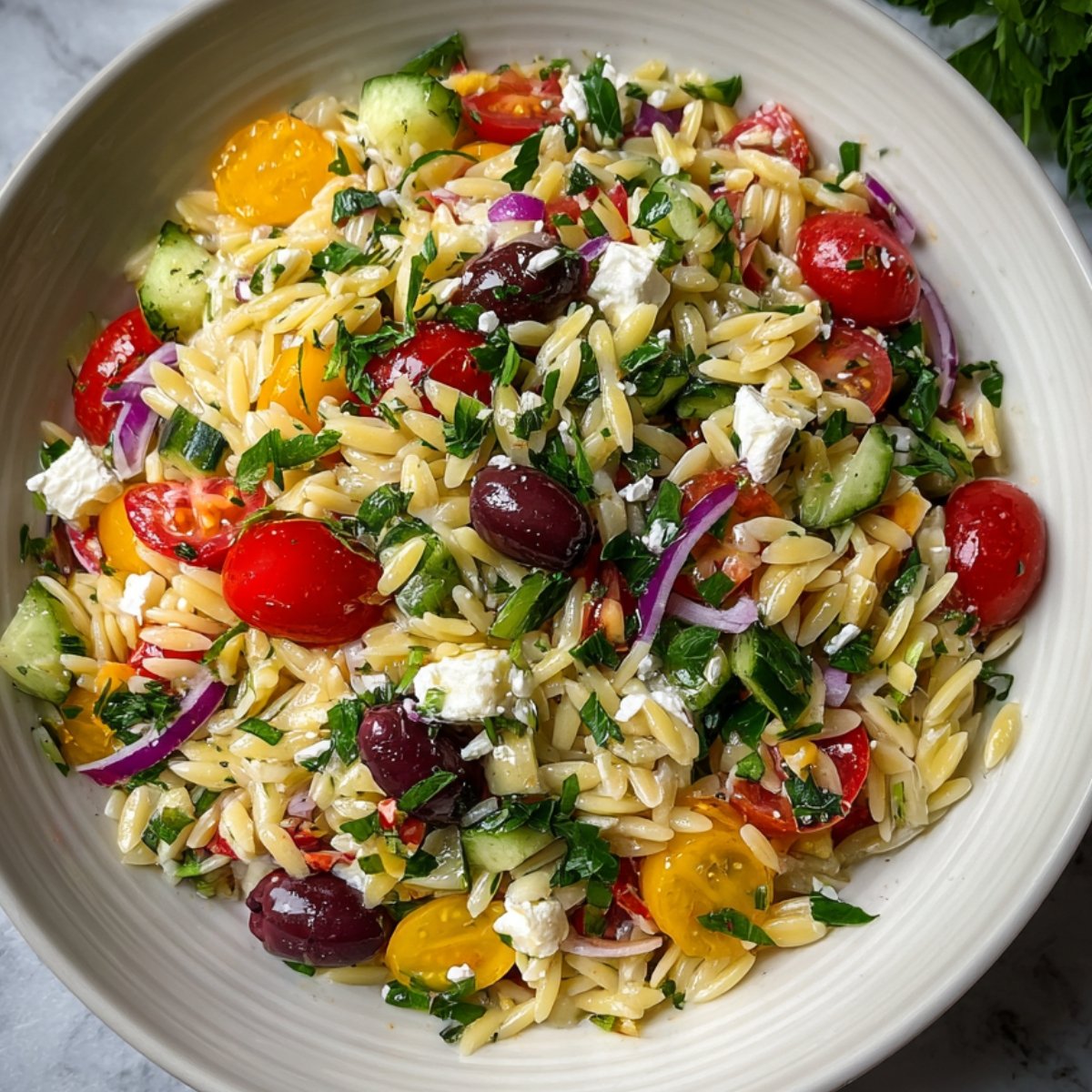 Close‑up overhead shot of finished Greek orzo salad in a white bowl, showing tender orzo mixed with cherry tomatoes, cucumber, red onion, parsley, Kalamata olives, and crumbled feta, all lightly coated in lemon‑olive oil dressing.”