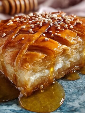 A slice of golden, flaky pastry drizzled with Honey Pie and topped with sesame seeds, served on a blue ceramic plate with a honey dipper in the background.