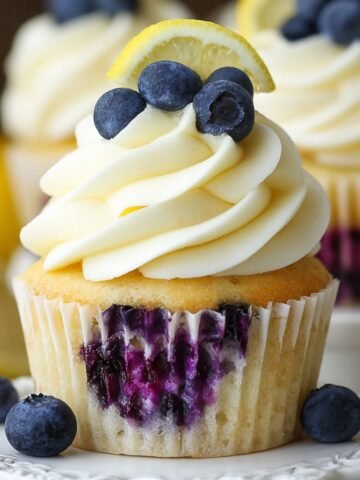A vanilla Lemon Blueberry Cupcakes studded with juicy blueberries, topped with swirled lemon‑cream cheese frosting, three fresh blueberries, and a thin lemon wedge, photographed against a softly blurred background.