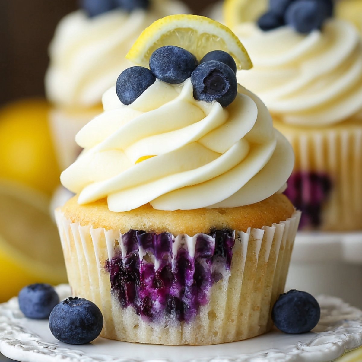 A vanilla Lemon Blueberry Cupcakes studded with juicy blueberries, topped with swirled lemon‑cream cheese frosting, three fresh blueberries, and a thin lemon wedge, photographed against a softly blurred background.