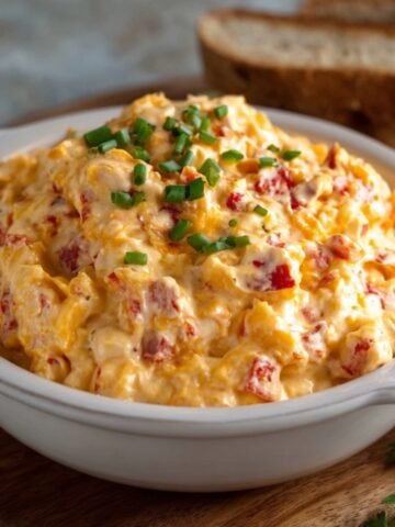 A creamy, homemade Pimento Cheese Recipe spread served in a white ceramic bowl, topped with chopped chives. The spread features a mix of shredded cheddar cheese, diced red pimentos, and mayonnaise, placed on a wooden board with slices of bread in the background.