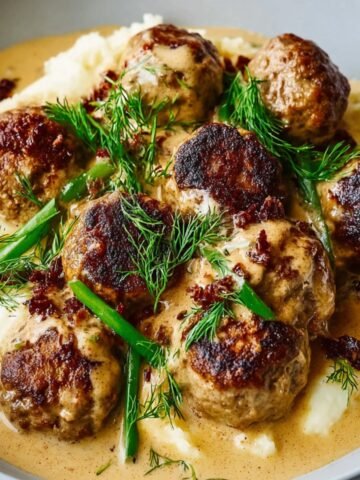 Swedish meatball recipe served in a creamy brown gravy over mashed potatoes, topped with fresh dill and chopped green onions.