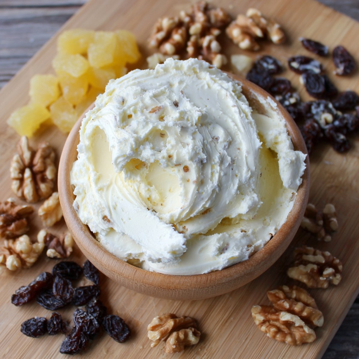 A wooden bowl piled high with swirled cream cheese frosting, surrounded by walnuts, dark raisins, and small cubes of candied pineapple on a wooden cutting board.