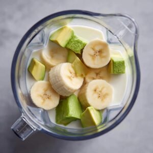 Close‑up of a tall glass filled with green avocado smoothie garnished with sesame seeds, with avocado halves and lime wedges in the background