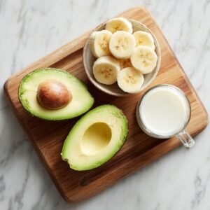 Wooden cutting board on a marble surface holding a halved avocado, a bowl of banana slices, and a glass of milk.