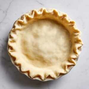 Overhead view of a round pie dish lined with raw, rolled‑out pastry dough. The edges are neatly crimped around the rim, and the pale dough sits on a white marble surface ready for filling.