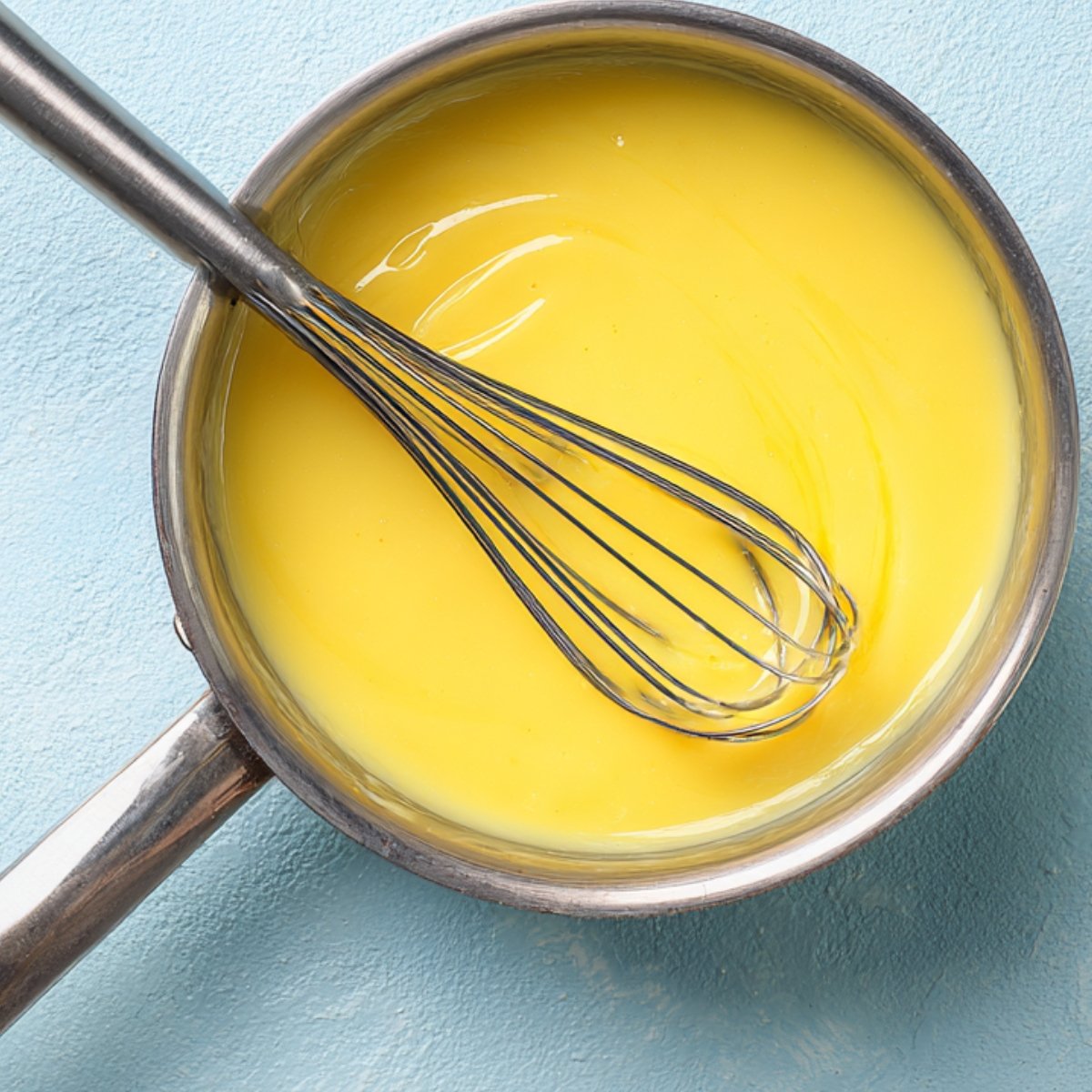 Stainless saucepan of smooth lemon curd with a whisk resting inside on a pale blue background.