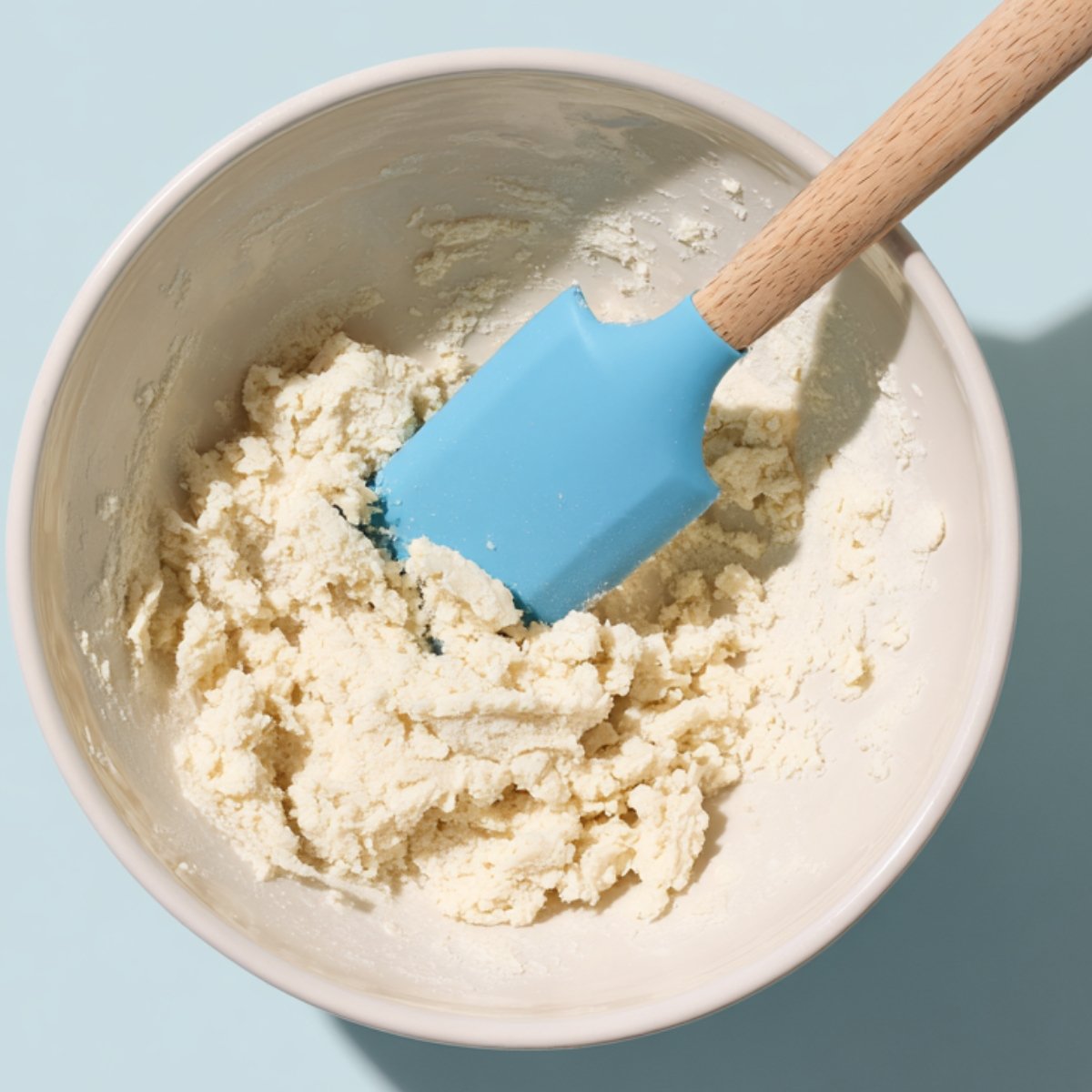 White mixing bowl of crumbly tart dough with a bright blue silicone spatula on a light blue surface.