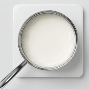 Overhead view of a stainless‑steel saucepan on a white induction cooktop, filled with plain whole milk heating steadily, the surface smooth and lightly steaming.