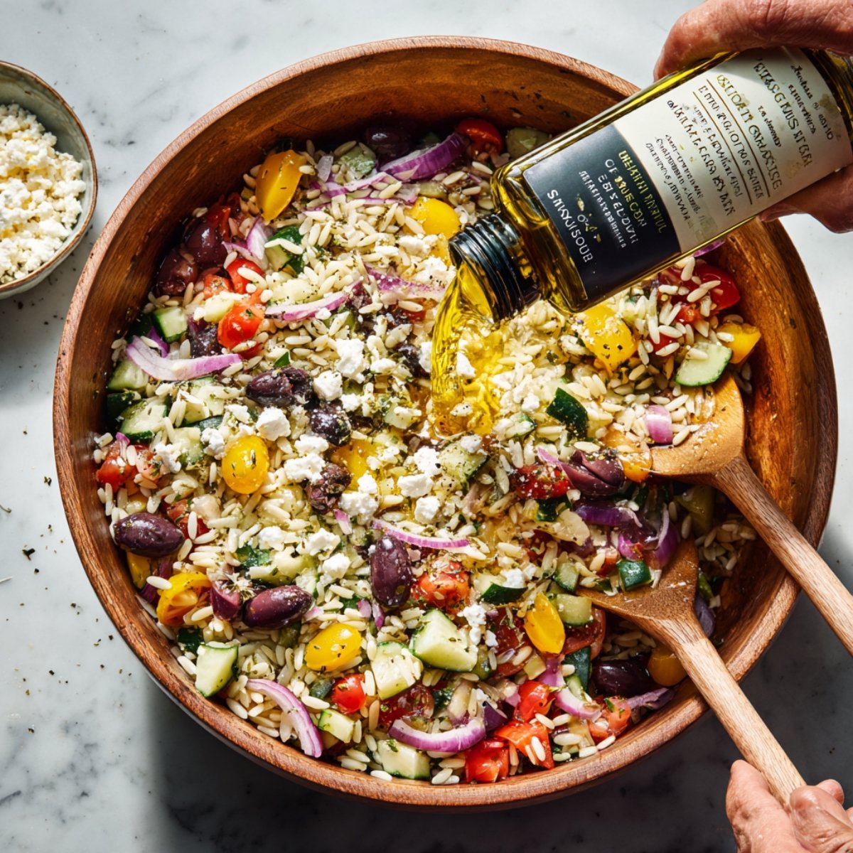 High‑angle photograph of a wooden salad bowl filled with orzo, tomatoes, cucumber, olives, red onion, and feta as golden olive oil is poured over it; two wooden servers rest inside the bowl