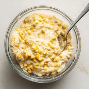 Overhead view of a clear mixing bowl half‑filled with creamy corn fritter batter studded with whole kernels, a silver fork resting inside, on a white marble countertop