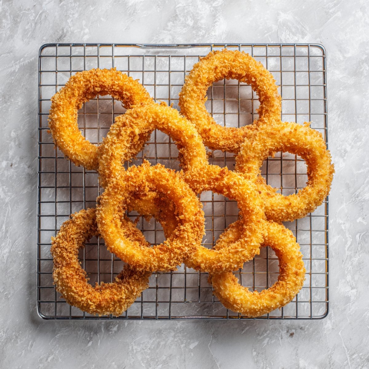 Breaded onion rings arranged on a wire rack over a light gray surface, ready to fry.