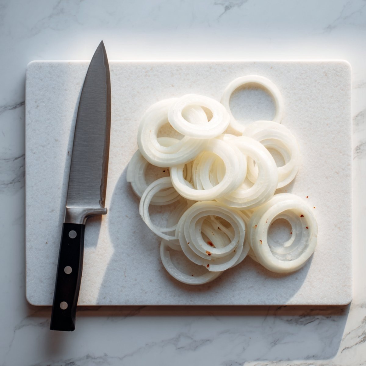 Thick onion rings and a chef’s knife on a white cutting board over a marble surface.