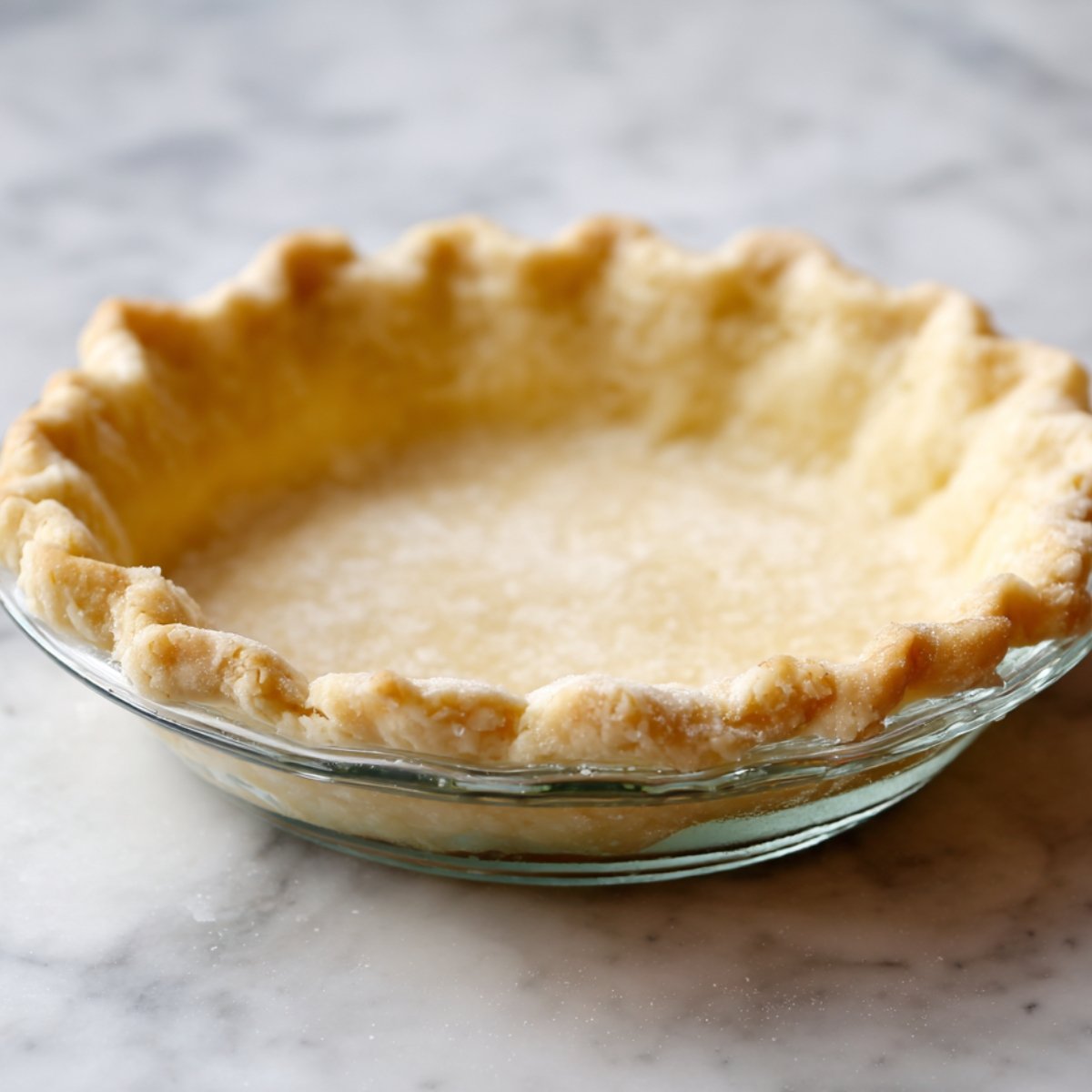 Prebaked pie crust with crimped edges in a clear glass dish, lightly golden and ready for filling.