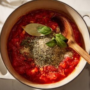 Top‑down view inside a white enamel Dutch oven on a marble countertop, showing bright red crushed tomatoes bubbling around a bay leaf and a heap of dried oregano and basil, with a wooden spoon resting at the rim.