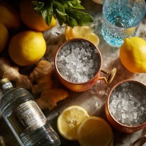 A flat lay of copper mugs filled with crushed ice, surrounded by lemons, fresh ginger, mint, and a bottle of vodka on a marble surface.