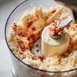 A high-quality photo showing a food processor filled with finely ground cheese and red pimentos. A spoon is scooping out a portion, showing the blended texture ready for forming or mixing.