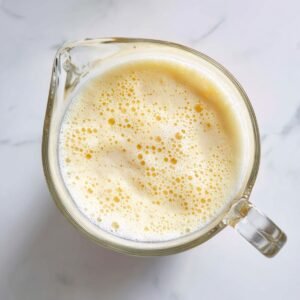 Overhead shot of a clear glass pitcher filled with frothy pale-yellow Piña Colada mixture, bubbles visible on the surface, set on a white marble countertop under diffuse daylight.