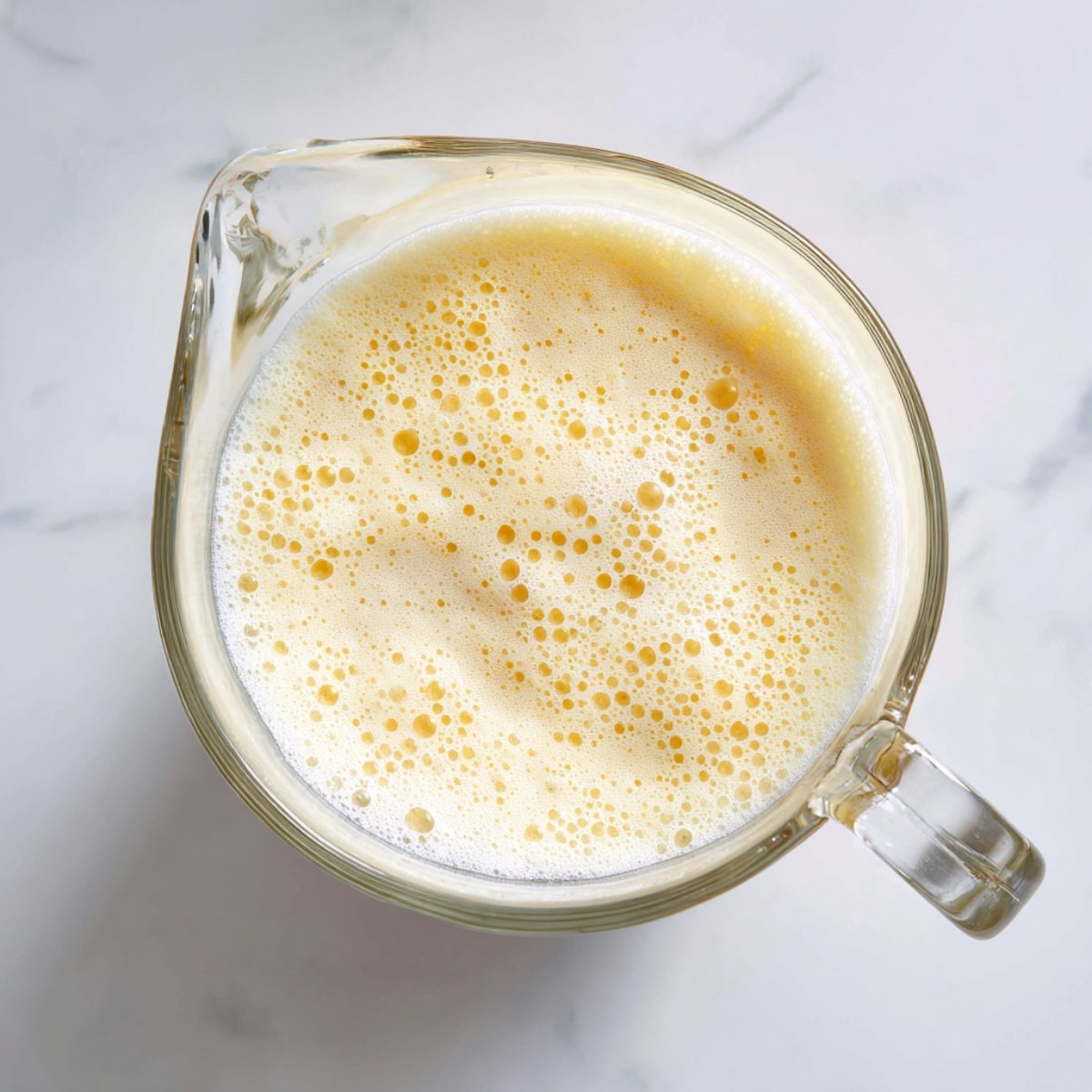 Overhead shot of a clear glass pitcher filled with frothy pale-yellow Piña Colada mixture, bubbles visible on the surface, set on a white marble countertop under diffuse daylight.