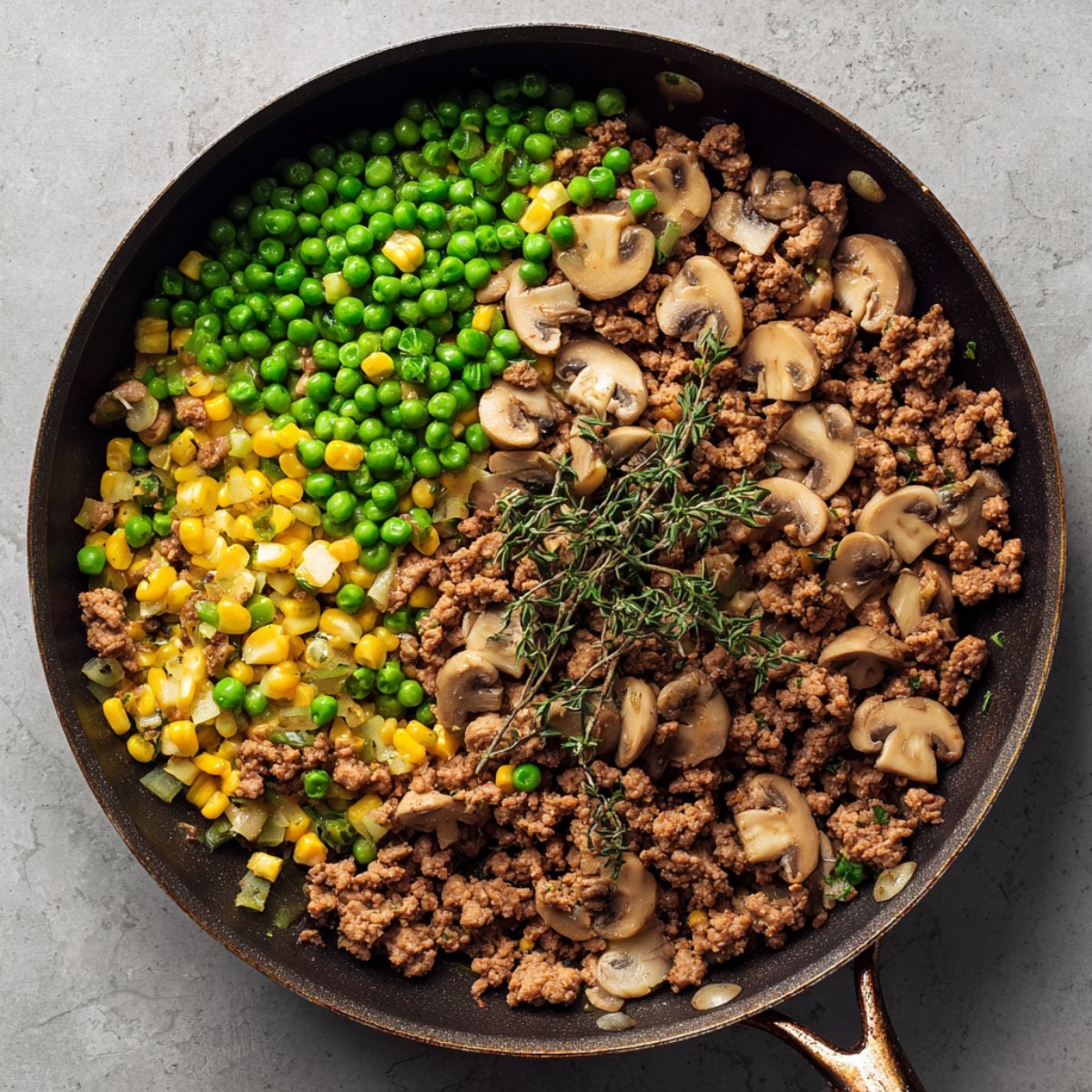 A top‑down view of a black skillet containing browned ground lamb mixed with bright green peas, yellow corn kernels, and sliced cremini mushrooms, topped with a few sprigs of fresh thyme.