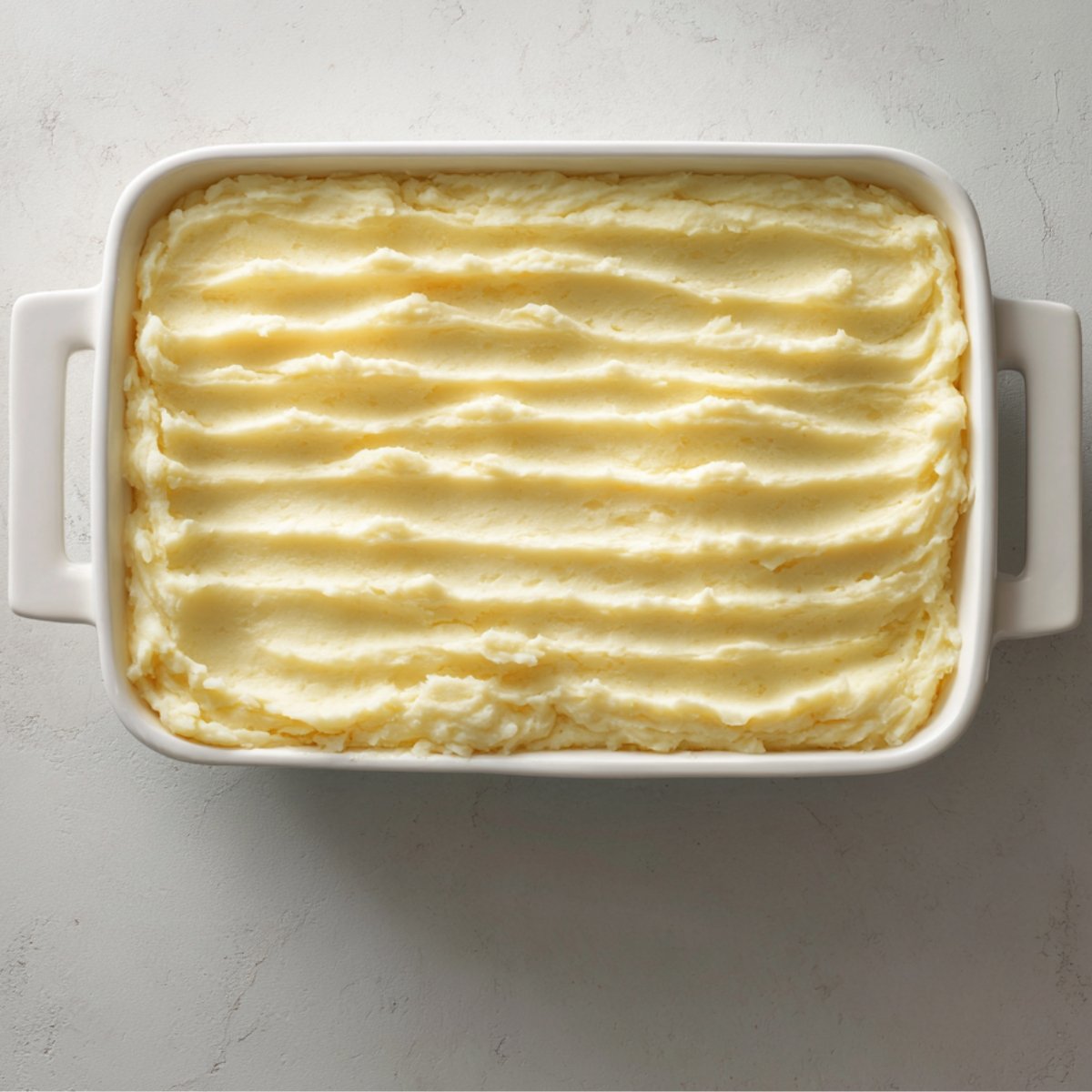 An overhead shot of the same white baking dish before baking: an even layer of pale mashed potatoes smoothed into parallel ridges covers the meat and vegetable layer below.