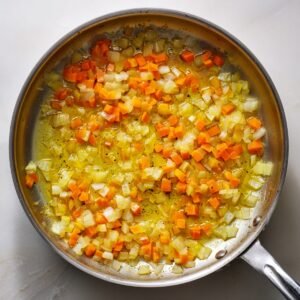 An overhead shot of a stainless‑steel skillet on a light surface, with diced yellow onion and carrot pieces sautéing in a thin sheen of oil until just softened.