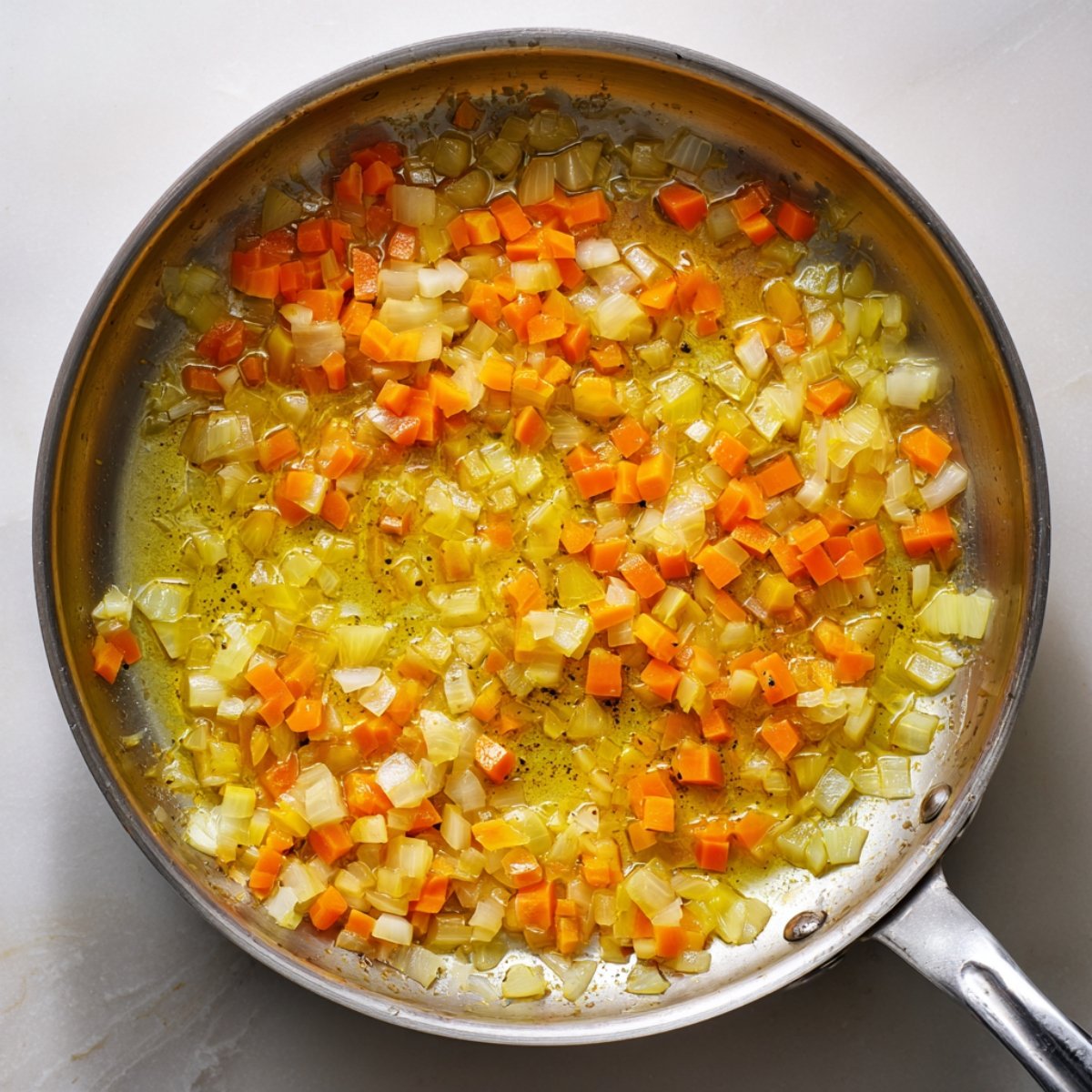 An overhead shot of a stainless‑steel skillet on a light surface, with diced yellow onion and carrot pieces sautéing in a thin sheen of oil until just softened.