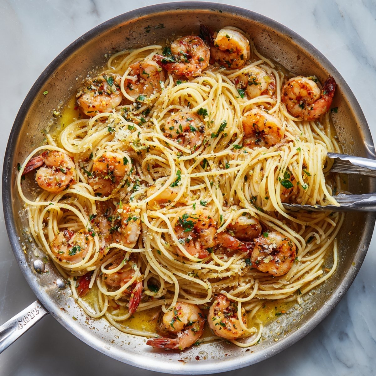 A stainless‑steel skillet filled with cooked spaghetti and sautéed shrimp, tossed in a garlic‑butter sauce and sprinkled with chopped parsley and grated cheese; a pair of tongs lifts a tangle of noodles.