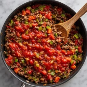 In a black skillet, cooked ground beef and diced vegetables are topped with a vibrant red tomato‑based sauce, ready to be folded in.