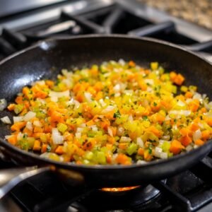 A black nonstick skillet on a gas burner with finely diced yellow onion, celery, and orange carrot pieces sautéing together.