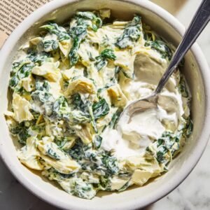 Top‑down view of a white ceramic bowl on a marble countertop, half‑filled with chopped spinach and artichoke hearts in creamy sauce, a stainless‑steel spoon resting inside
