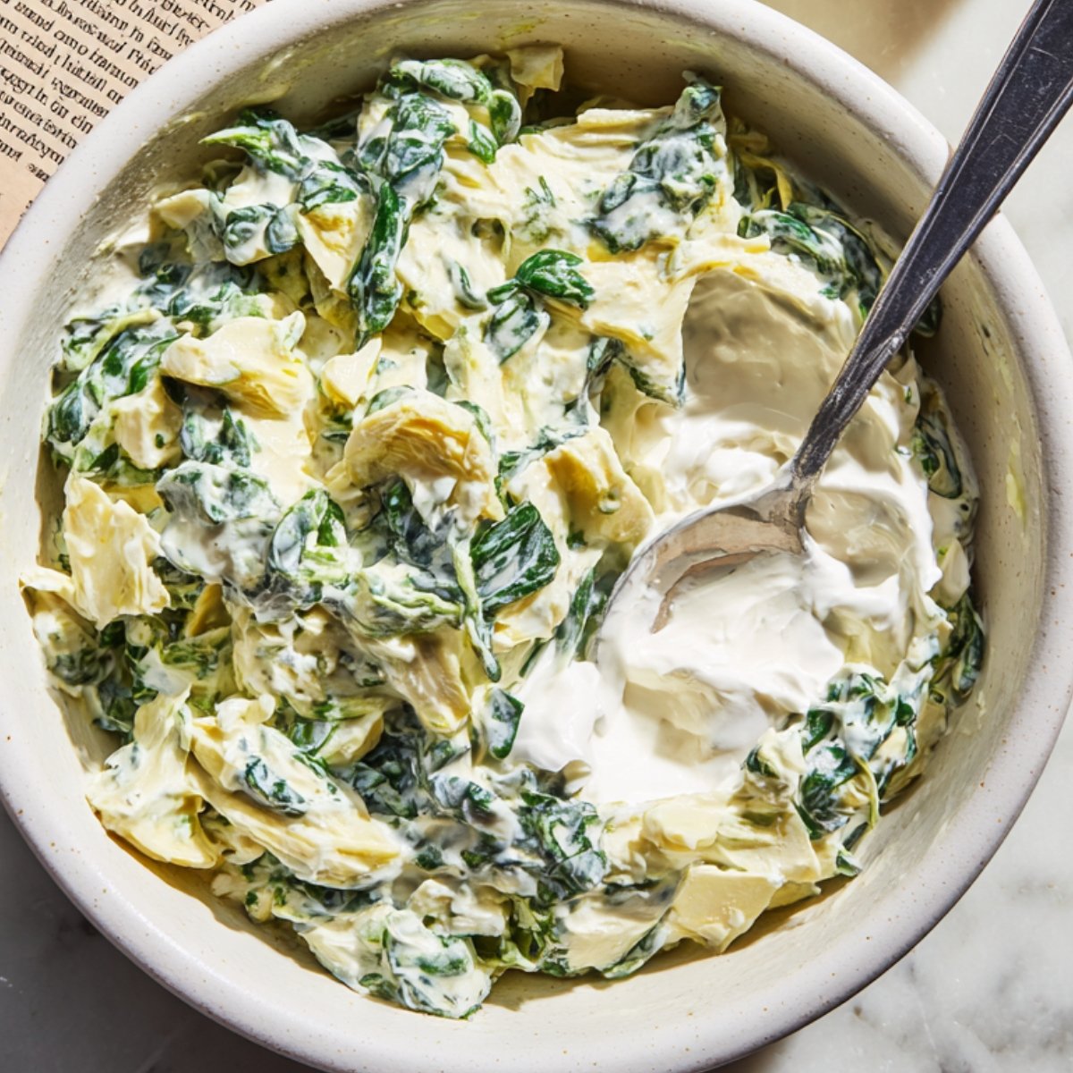 Top‑down view of a white ceramic bowl on a marble countertop, half‑filled with chopped spinach and artichoke hearts in creamy sauce, a stainless‑steel spoon resting inside
