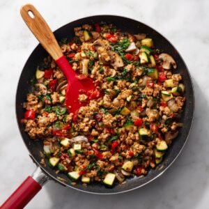 Top-down shot of a black sauté pan on marble, packed with browned ground meat, zucchini, mushrooms, spinach, onions, and red peppers, with a red spatula resting inside.