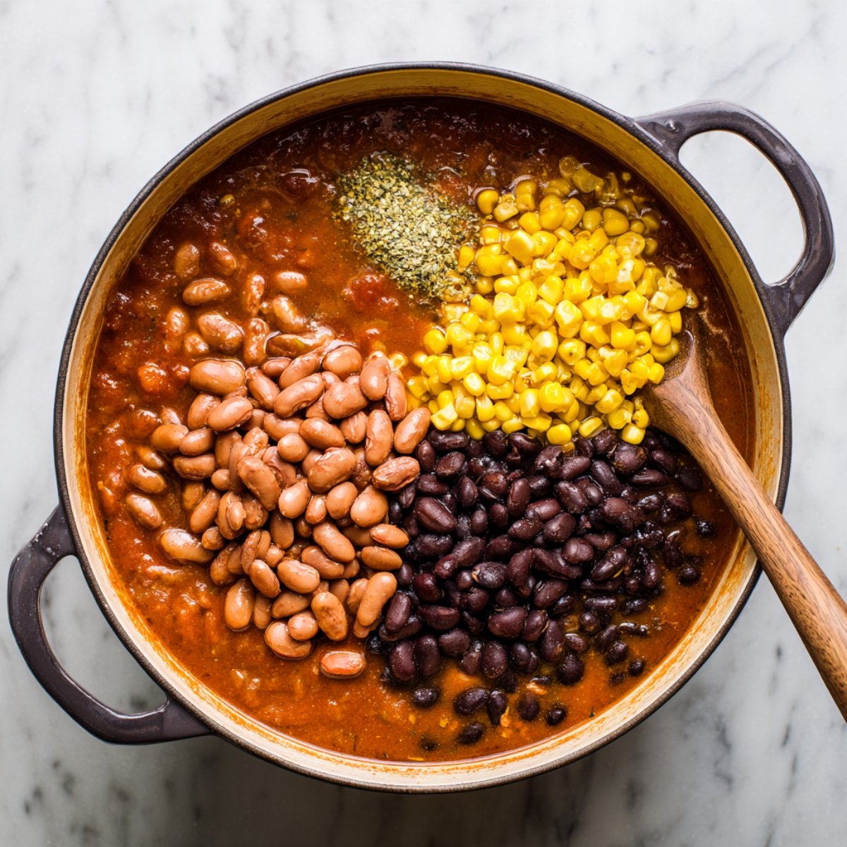 Top-down shot of a white pot on a marble counter, showing three separate heaps of ingredients in broth: yellow corn kernels in the upper half, light brown pinto beans at lower left, and black beans at lower right, before stirring.