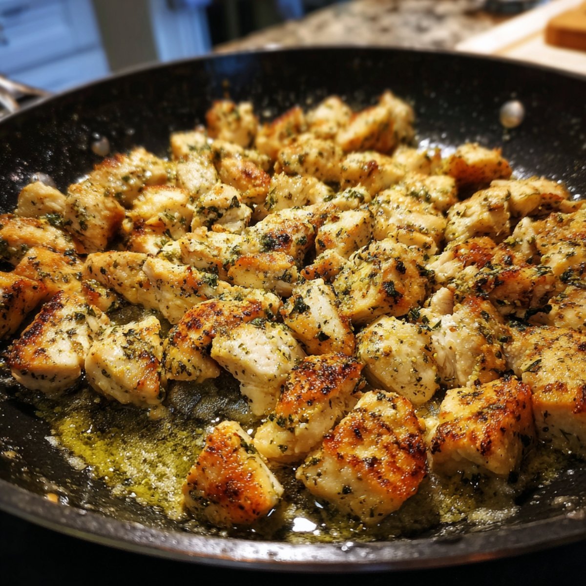 Close‑up side view of a large skillet with bite‑sized chicken cubes sizzling in oil, each piece browned to a crisp golden crust beneath kitchen lighting.