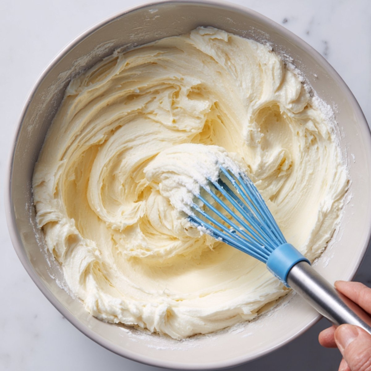 A mixing bowl of whipped cream‑cheese filling; a hand holds a blue silicone whisk, scraping and stirring the thick, creamy mixture until smooth.