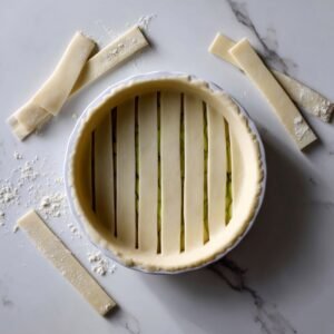 Overhead of a golden-brown lattice-topped apple pie resting in a red ceramic dish on a wire cooling rack over a marble countertop, the sugar-speckled crust crisp and flaky, with tender apple filling peeking through the woven pattern.