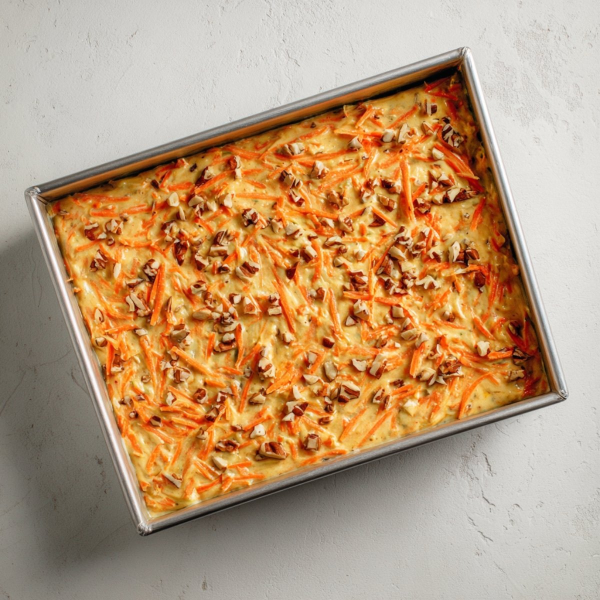 Angled overhead image of a rectangular metal baking pan entirely filled with unbaked carrot cake batter, showing visible shreds of carrot and pecan pieces on the surface.