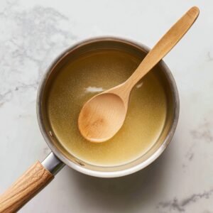 Overhead view of a saucepan on a marble counter, containing golden simple syrup being stirred by a wooden spoon until fully dissolved.
