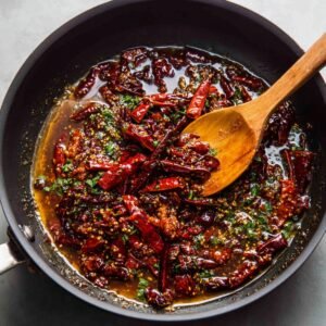 A bright marble countertop displaying small ceramic bowls of fresh orange juice, grated orange zest, soy sauce, granulated sugar, olive oil, minced garlic, red pepper flakes, sesame seeds, thinly sliced scallion greens, whole garlic bulb, and a knob of ginger, all neatly arranged in a flat lay.