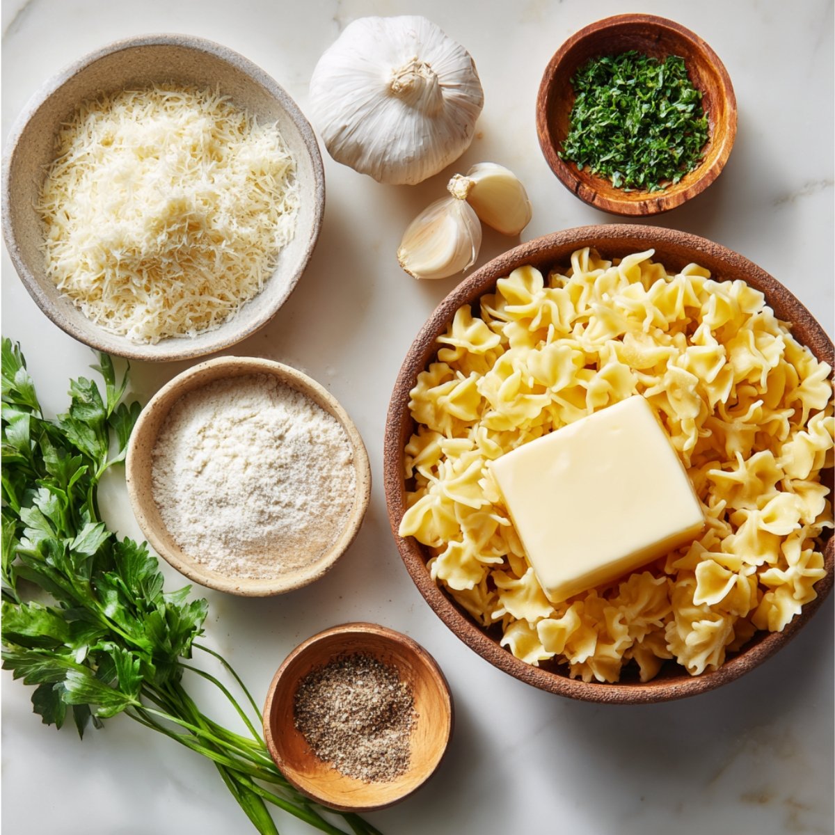 Overhead view of ingredients for homemade Alfredo sauce: a wooden bowl of uncooked egg noodles topped with a square of butter, bowls of freshly grated Parmesan and grated Pecorino cheese, a head of garlic with cloves, a small bowl of chopped parsley, and a small wooden dish of cracked black pepper on a marble surface.