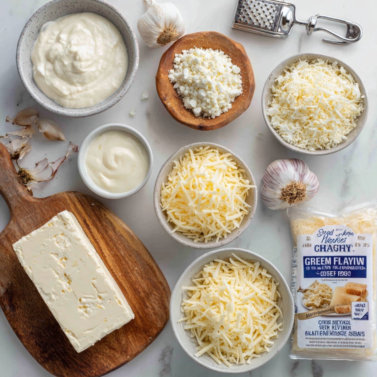 Overhead flat‑lay of creamy dip ingredients on a white surface: a bowl of sour cream, a small dish of mayonnaise, a wooden bowl of grated Parmesan, a bowl of shredded mozzarella, a bowl of shredded sharp cheddar, a head of garlic, and a stainless‑steel garlic press.
