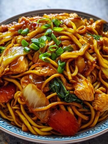 A savory dish of stir-fried noodles with Chicken Chow Mein, tossed in a rich soy-based sauce, mixed with vegetables like onions, carrots, and leafy greens, and garnished with chopped green onions. Served in a patterned bowl.