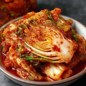 Bowl of freshly made Fresh Kimchi featuring napa cabbage in a spicy red pepper paste, with a jar of kimchi in the background on a dark surface.
