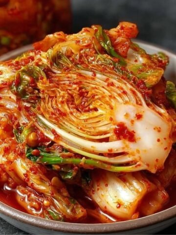 Bowl of freshly made Fresh Kimchi featuring napa cabbage in a spicy red pepper paste, with a jar of kimchi in the background on a dark surface.